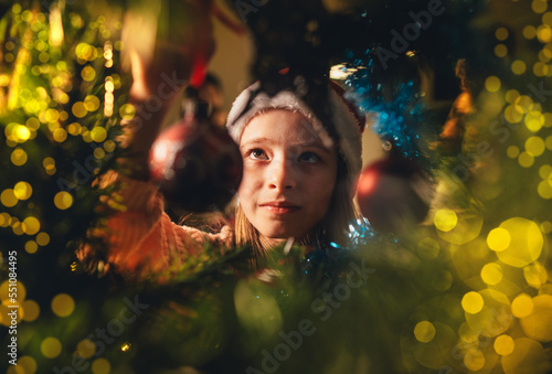 Cute little girl in red Santa hat hanging a magic tree decoration ball on the Christmas fir-tree on fairy tale x-mass wishes night. Charming childhood and winter holidays concept inside branches image