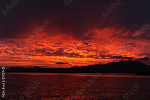 Red, pink, orange and violet sunrise at the seaside during dawn with clouds in the sky and mountains on the shore of a port © piotrmilewski