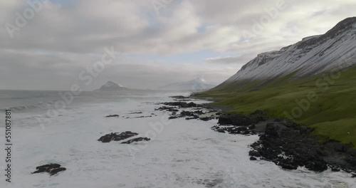 aerial movement over rocky shore of greenland sea at coast of iceland with green grass and moutnains with snow  in wesfjords