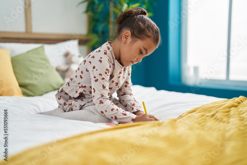 Adorable hispanic girl drawing on notebook sitting on bed at bedroom
