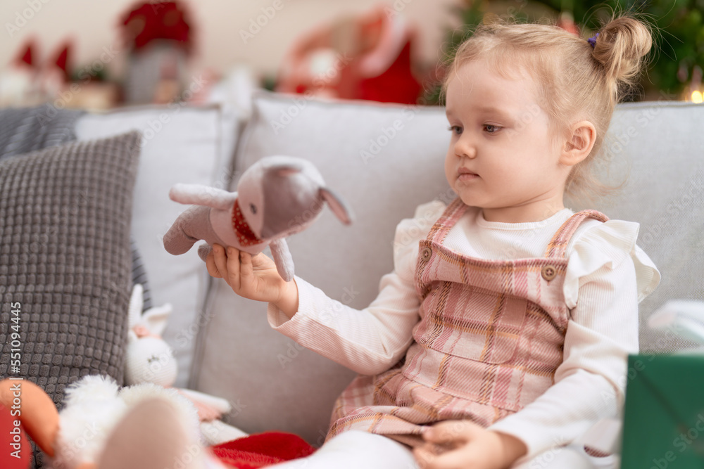 Fototapeta premium Adorable caucasian girl holding toy sitting on sofa by christmas tree at home
