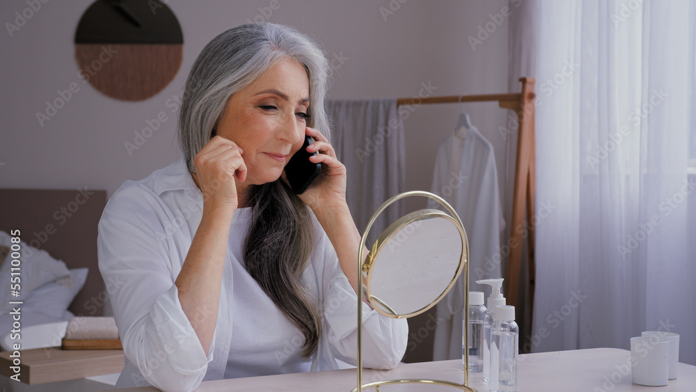 Senior gray-haired old 60s woman lady talking mobile phone talk conversation looking at mirror reflection buying ordering face cream by smartphone enjoy wrinkled beautiful aging perfect smooth skin Senior gray-haired old 60s woman lady talking mobile phone talk conversation looking at mirror reflection buying ordering face cream by smartphone enjoy wrinkled beautiful aging perfect smooth skin