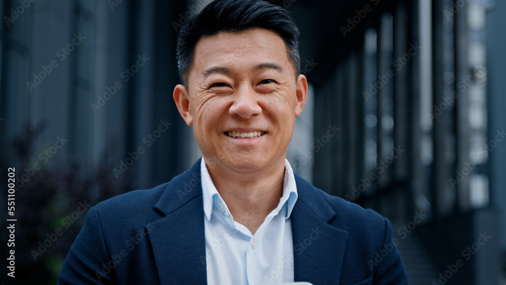 Glad Asian 40s businessman with smartphone texting browsing smiling looking at camera standing outdoors in city. Middle-aged man CEO has business chat using mobile phone check news in social net Glad Asian 40s businessman with smartphone texting browsing smiling looking at camera standing outdoors in city. Middle-aged man CEO has business chat using mobile phone check news in social net