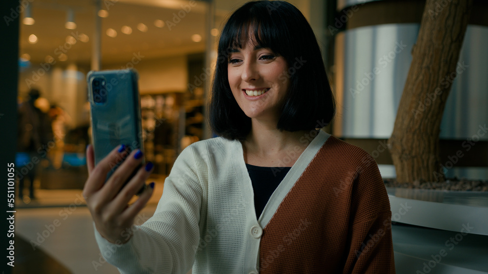 Smiling positive talking to smartphone webcam woman has video call indoors in shopping mall waving hand hello hi greeting girl speak phone conference distant chat virtual meeting talk remote speaking Smiling positive talking to smartphone webcam woman has video call indoors in shopping mall waving hand hello hi greeting girl speak phone conference distant chat virtual meeting talk remote speaking