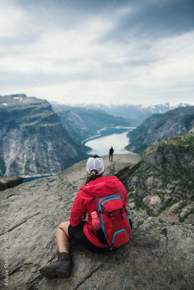 A young tourist woman in red jacket sitting on the rock enjoying the beautiful view on norwegian very famous place called Trolltunga. Summer hiking in norway nature. Breathtaking.Relax mood