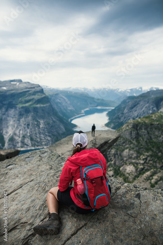 A young tourist woman in red jacket sitting on the rock enjoying the beautiful view on norwegian very famous place called Trolltunga. Summer hiking in norway nature. Breathtaking.Relax mood