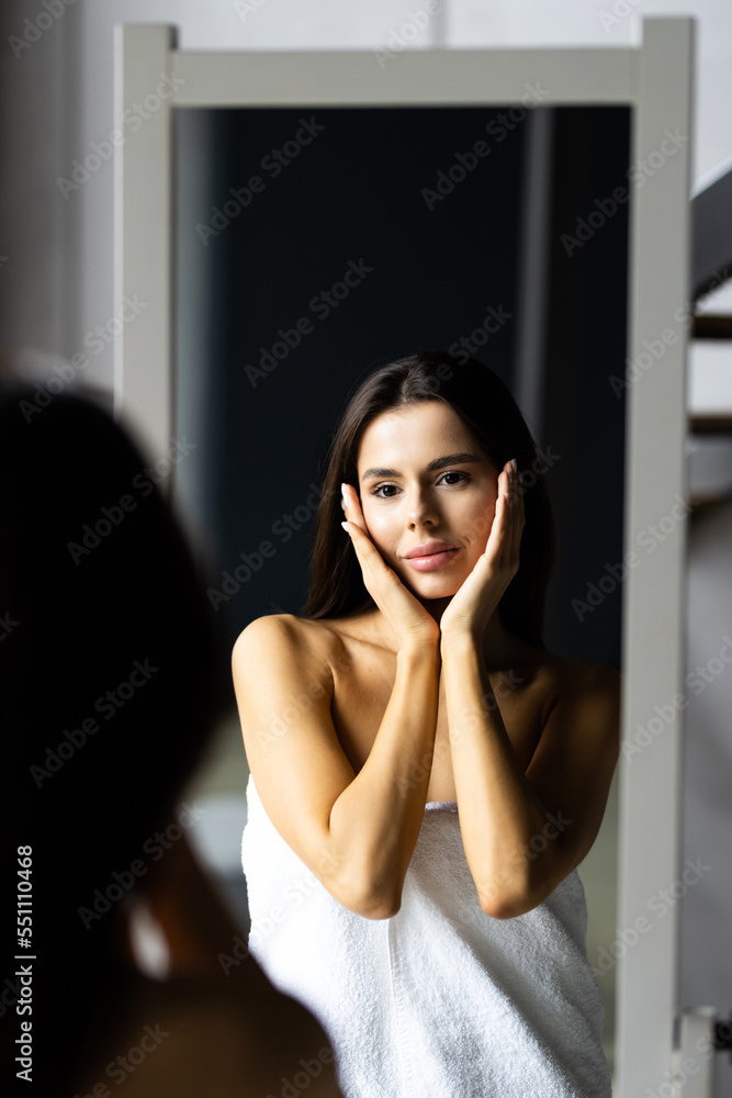 Self-Care Concept. Portrait of attractive young female looking at mirror. Beautiful Woman Wearing White Silk Robe Touching Soft Skin On Face And Smiling, Enjoying Her Reflection, Selective Focus