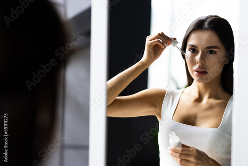 Cheerful woman standing in front of mirror at home, using serum for hydrating skin at summer
