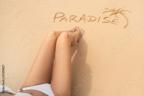 Word paradise and one palm on the sandy beach near the women's legs. Summer vacation concept. Realax. photo