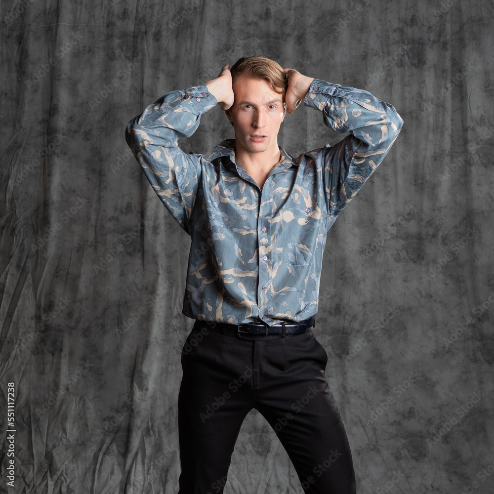 Stylish elegant young man in a blue silk shirt posing in the studio on a gray fabric background