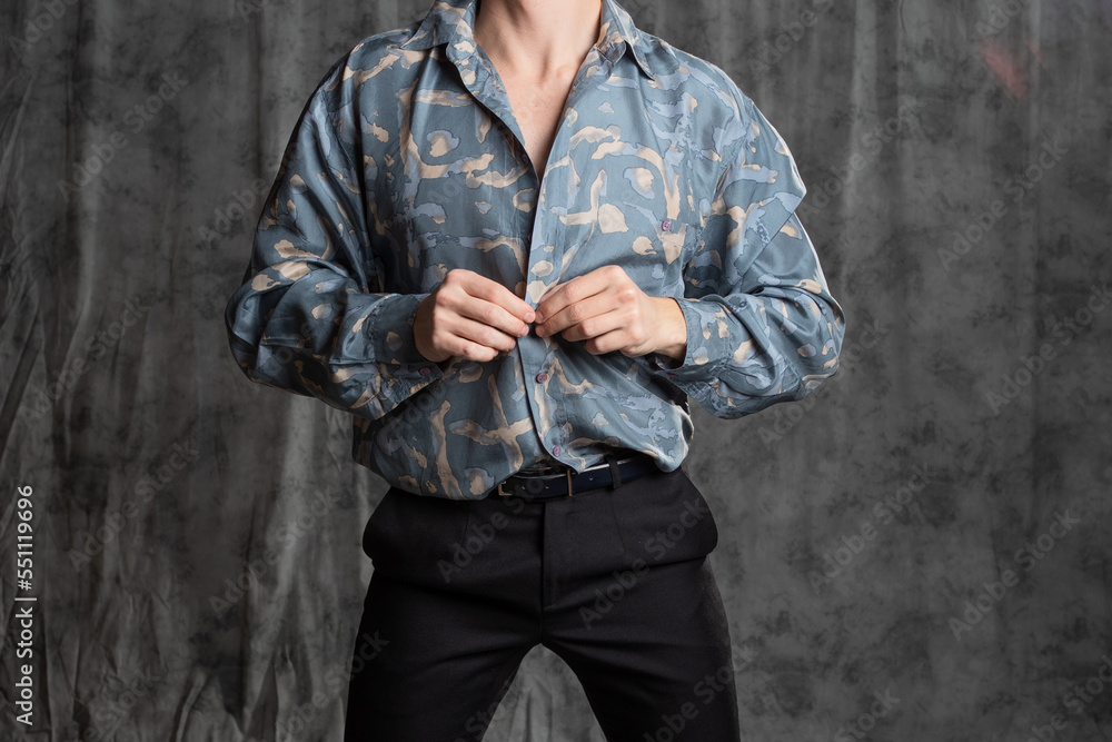 Stylish elegant young man in a blue silk shirt posing in the studio on a gray fabric background. sexy pose. Unbuttoning the shirt on the chest, close-up Stylish elegant young man in a blue silk shirt posing in the studio on a gray fabric background. sexy pose. Unbuttoning the shirt on the chest, close-up