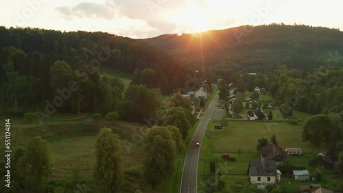 Cars drive on road passing through small village in highland of Europe. Sun sets behind giant forestry mountains aerial view photo