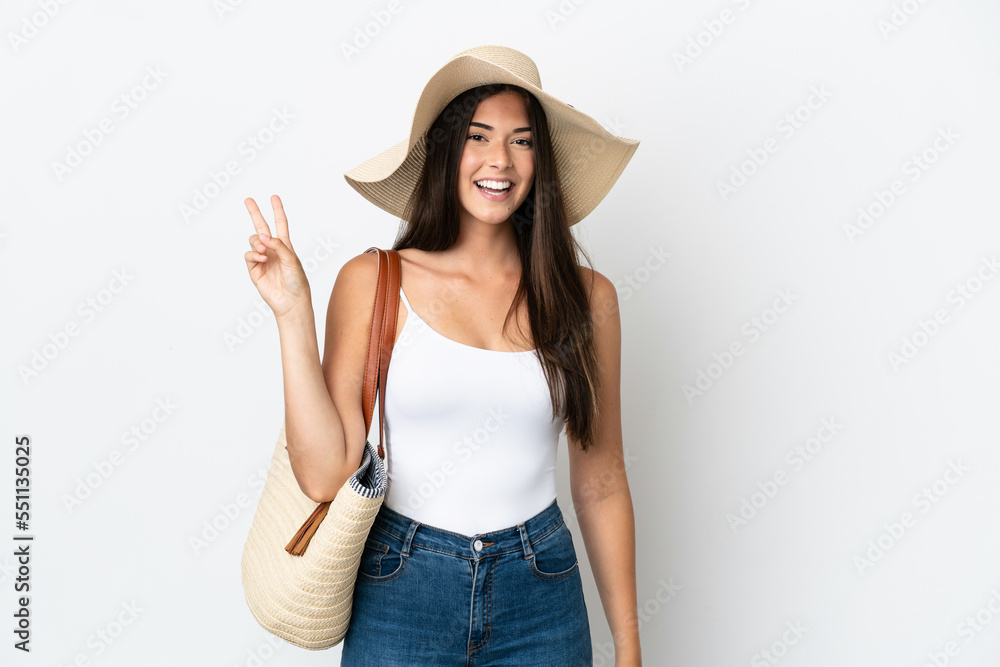Young Brazilian woman with Pamela holding a beach bag isolated on white background smiling and showing victory sign Young Brazilian woman with Pamela holding a beach bag isolated on white background smiling and showing victory sign