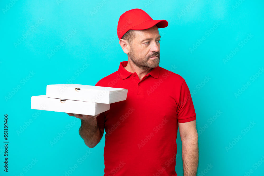 Pizza delivery man with work uniform picking up pizza boxes isolated on blue background looking to the side Pizza delivery man with work uniform picking up pizza boxes isolated on blue background looking to the side