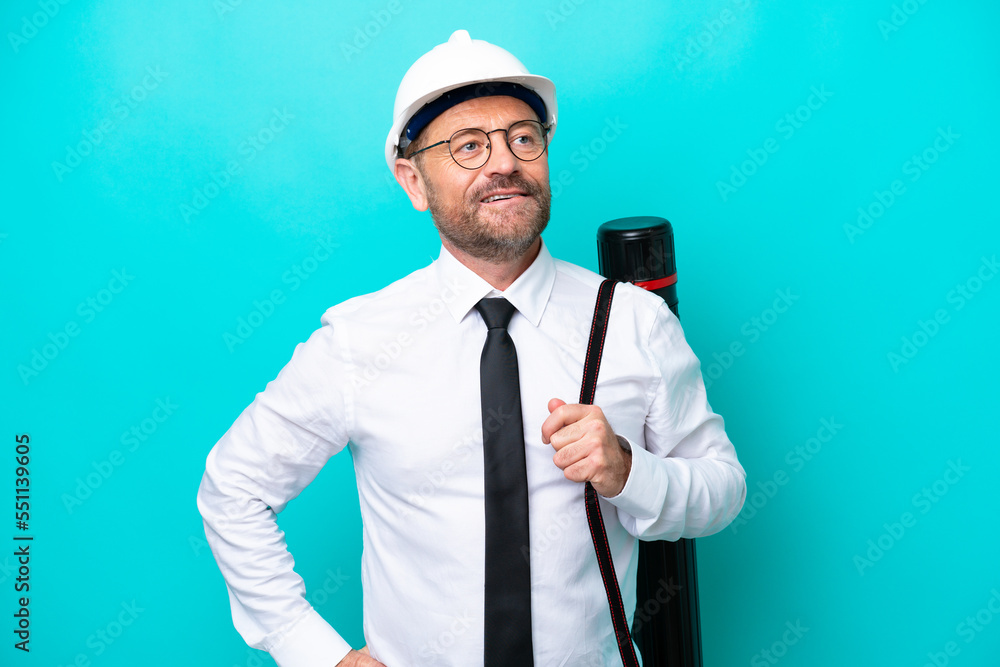 Middle age architect man with helmet and holding blueprints isolated on blue background posing with arms at hip and smiling Middle age architect man with helmet and holding blueprints isolated on blue background posing with arms at hip and smiling