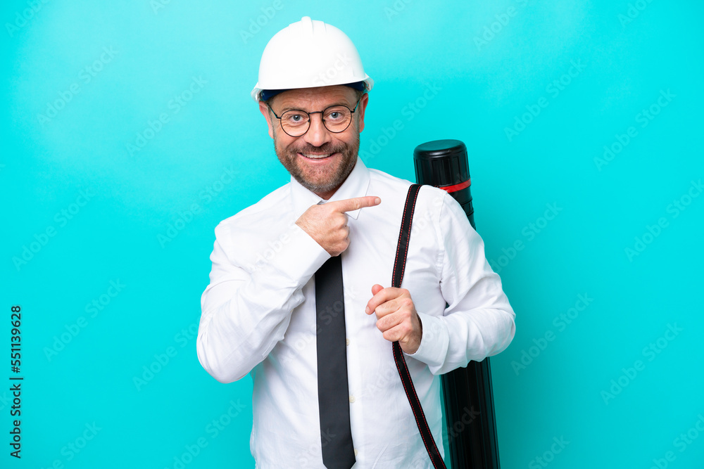 Middle age architect man with helmet and holding blueprints isolated on blue background pointing to the side to present a product Middle age architect man with helmet and holding blueprints isolated on blue background pointing to the side to present a product
