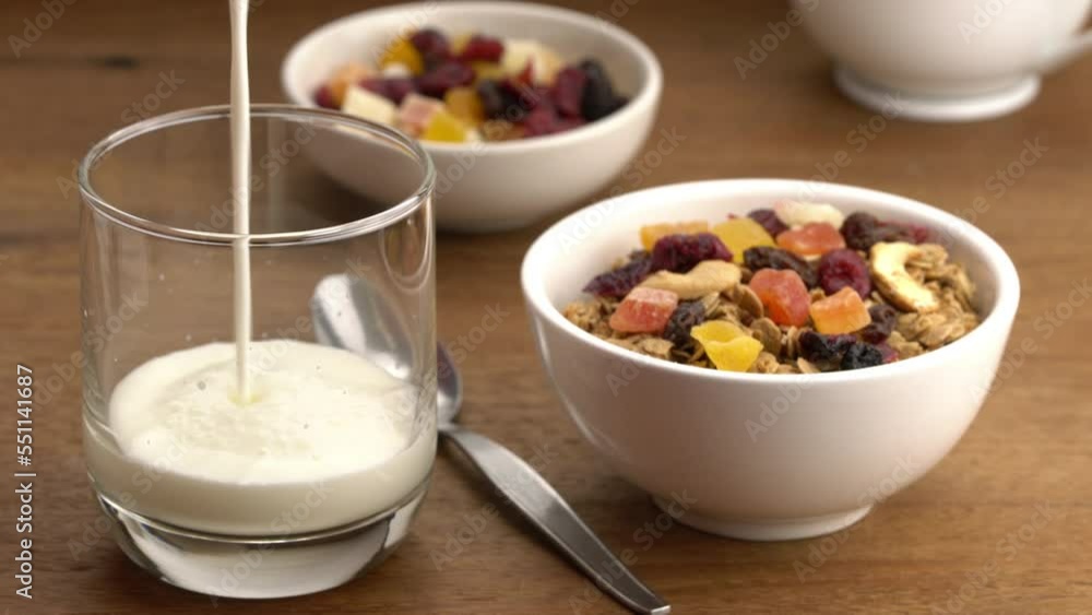 Pouring fresh milk into transparent glass with crunchy muesli cereal breakfast and various dry preserved sweet tropical fruits in white ceramic bowl on wooden table. Pouring fresh milk into transparent glass with crunchy muesli cereal breakfast and various dry preserved sweet tropical fruits in white ceramic bowl on wooden table.