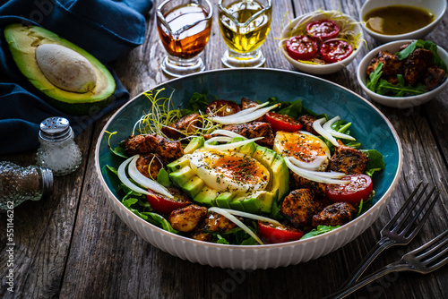 Tasty salad - fried chicken breast, avocado, boiled eggs, mini tomatoes and fresh green vegetables on wooden background 