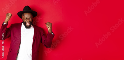 Successful Black man in suit saying yes, rejoicing of winning or achieving goal, raising hands up and screaming of joy, red background photo