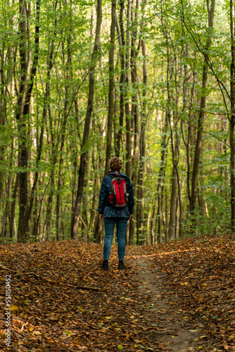 Autumn walk in the forest. A woman with a red backpack hiking on a forest trail in the fall. Green trees and dried leaves. Walking in forest. © Łukasz T.
