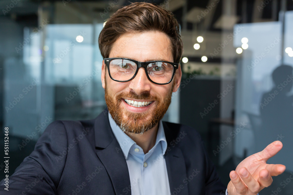 Businessman looks into smartphone camera and talks on video call, man uses phone for remote communication with friends and colleagues, investor boss in glasses works inside office.