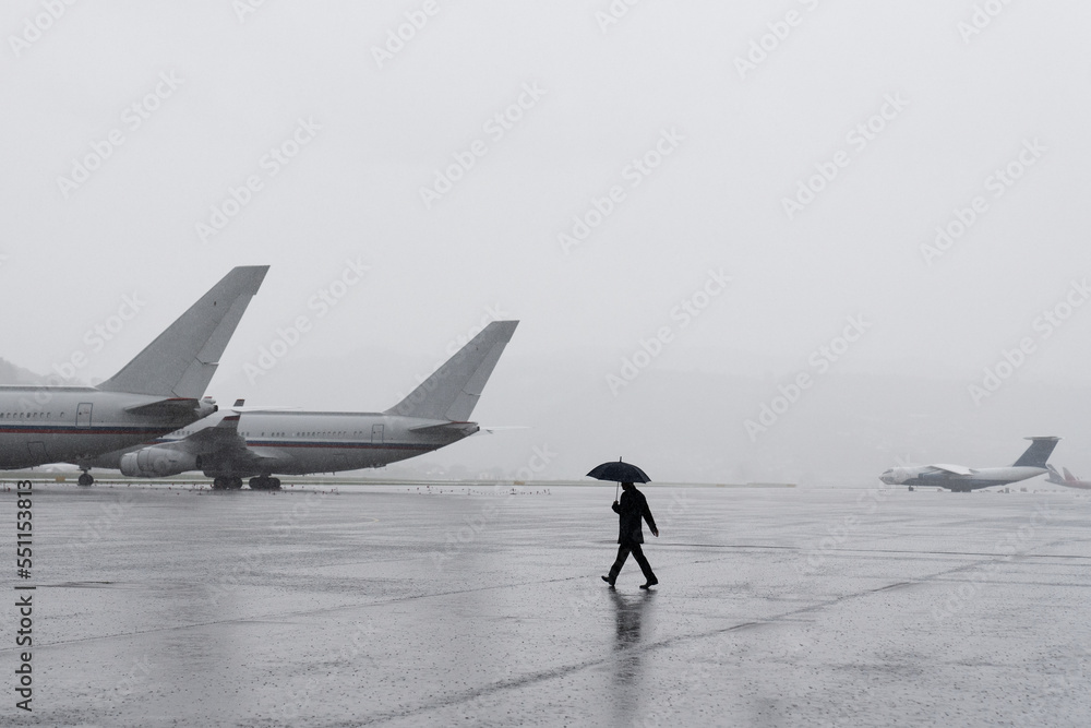 Naklejka premium A man under an umbrella at the airport. Desert airfield. Rainy weather at the airport. Large airfield with aircraft. The man goes to the plane.