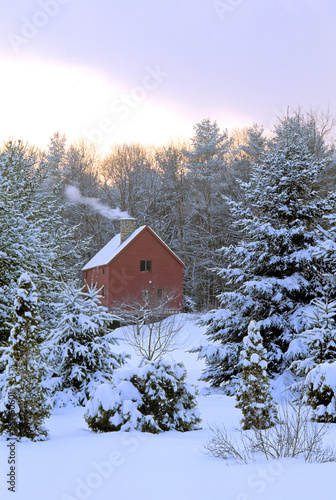 Tranquil New England winter scene following a departing snowstorm. Clearing sky, warm cozy home with smoke rising from stone chimney, and trees covered in fresh snow.