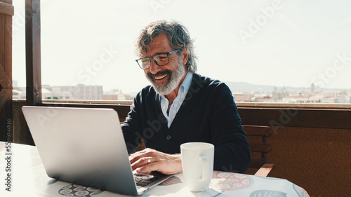 Happy middle aged man communicates remotely using laptop. Working from home office