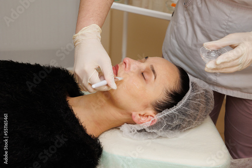 Close up face of woman face in white medical hat. Professional cosmetologist in whitemedical gloves apply cream with a tassel. Anti-aging treatment and face massage in cosmetology clinic.  photo