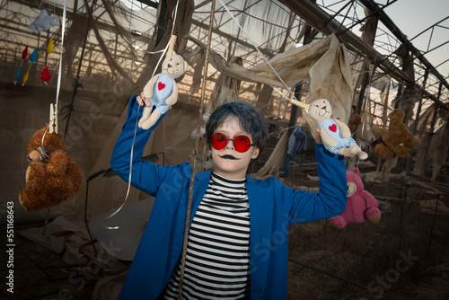 a blue-haired boy in red sunglasses, a scary smile in a blue jacket and makeup is having fun and fooling around in an abandoned greenhouse among hanging toys