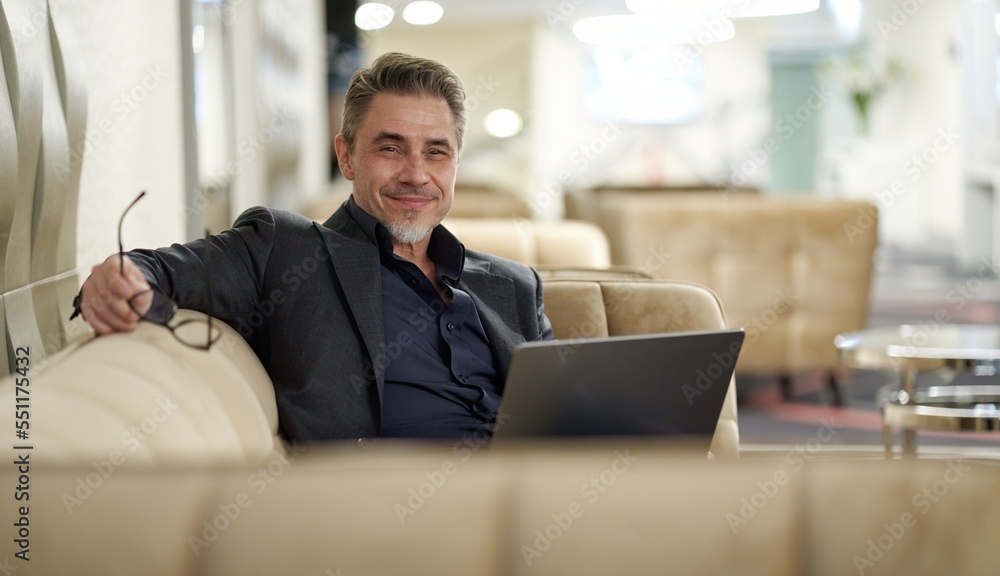 Businessman working in business lounge. Entrepreneur sitting on sofa in office lobby using laptop computer to remote work online. Happy middle aged, mid adult, mature age man smiling. Businessman working in business lounge. Entrepreneur sitting on sofa in office lobby using laptop computer to remote work online. Happy middle aged, mid adult, mature age man smiling.