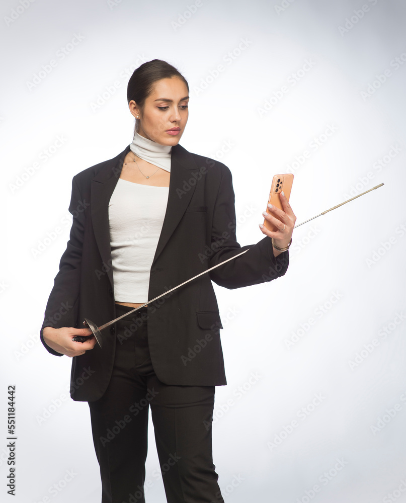 professional young woman with a rapier and a smartphone tablet. businesswoman in a business suit with a sword in her hand uses a tablet smartphone on a white background isolated. professional young woman with a rapier and a smartphone tablet. businesswoman in a business suit with a sword in her hand uses a tablet smartphone on a white background isolated.