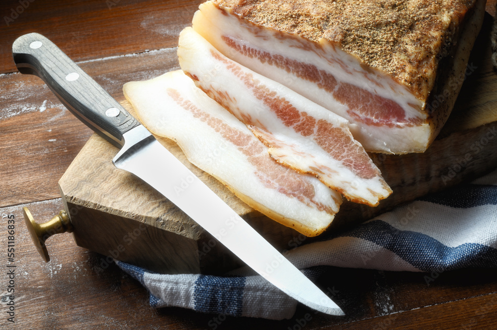 Bacon, guanciale, with cutting board and knife on wooden background, close-up.