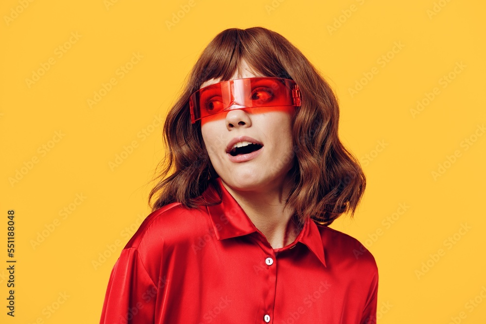 a stylish woman stands on a yellow background in a red shirt and glasses of an interesting shape smiling pleasantly at the camera