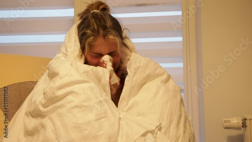 Sick woman in home room with dim light wrap up in warm white blanket and blow nose into handkerchief. Stuffy nose, snot.