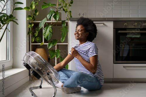 Young happy satisfied African American woman sitting on floor in kitchen in front of air cooler, black girl using fan to cool down at home. Overjoyed pleased female cooling herself with ventilator