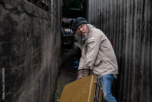 Asian poverty homeless man walk dragging large suitcase in narrow alley find for rest, poor man live in slump no job, pity aged person problem from financial crisis unemployment without family. © Rakchanok