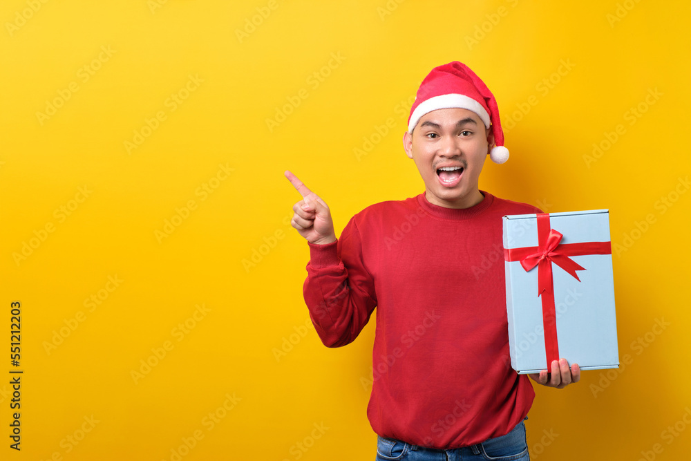 Surprised young Asian man in Santa hat holding gift box with ribbon, pointing finger aside at copy space on yellow studio background. celebration Christmas holiday and New Year concept Surprised young Asian man in Santa hat holding gift box with ribbon, pointing finger aside at copy space on yellow studio background. celebration Christmas holiday and New Year concept