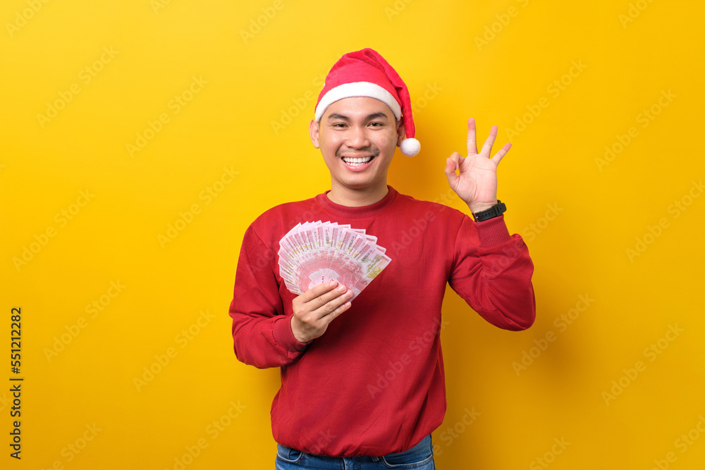 Cheerful young Asian man in Santa hat holding fan of cash money, showing okay gesture on yellow studio background. celebration Christmas holiday and New Year concept Cheerful young Asian man in Santa hat holding fan of cash money, showing okay gesture on yellow studio background. celebration Christmas holiday and New Year concept