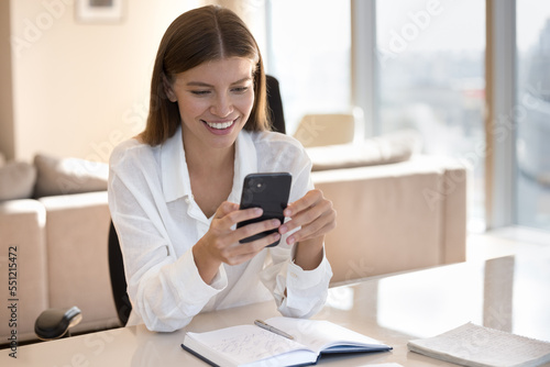 Happy busy young business woman using mobile phone, sitting at notebook, workplace table, chatting online, working from home office, using app, enjoying internet job communication