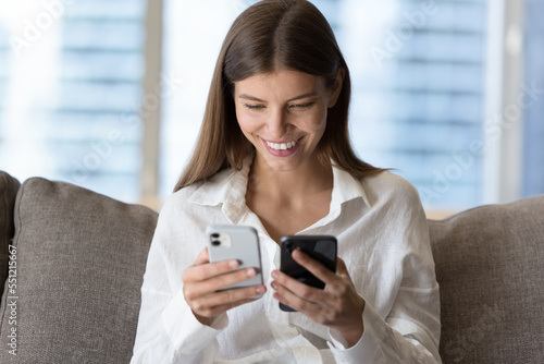 Happy young millennial Caucasian girl holding two mobile phones, using wireless connection technology, gadgets for data transfer, looking at screen, smiling, laughing, sitting on home sofa