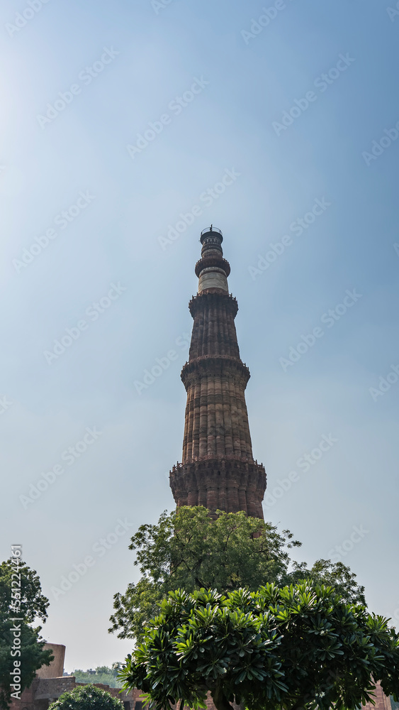 The world's tallest brick minaret Qutub Minar against the blue sky. Wavy red walls, several openwork balconies are visible. Lush green vegetation at the foot. India. Delhi