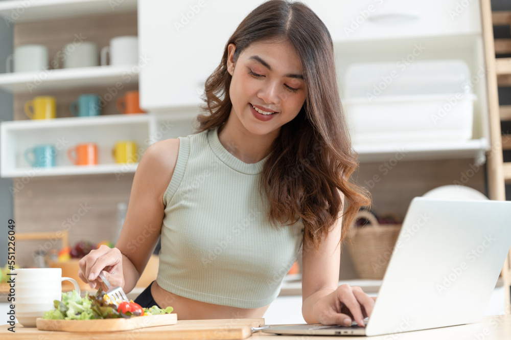 Happy Asia woman eating salad with see computer notebook in kitchen room Happy Asia woman eating salad with see computer notebook in kitchen room