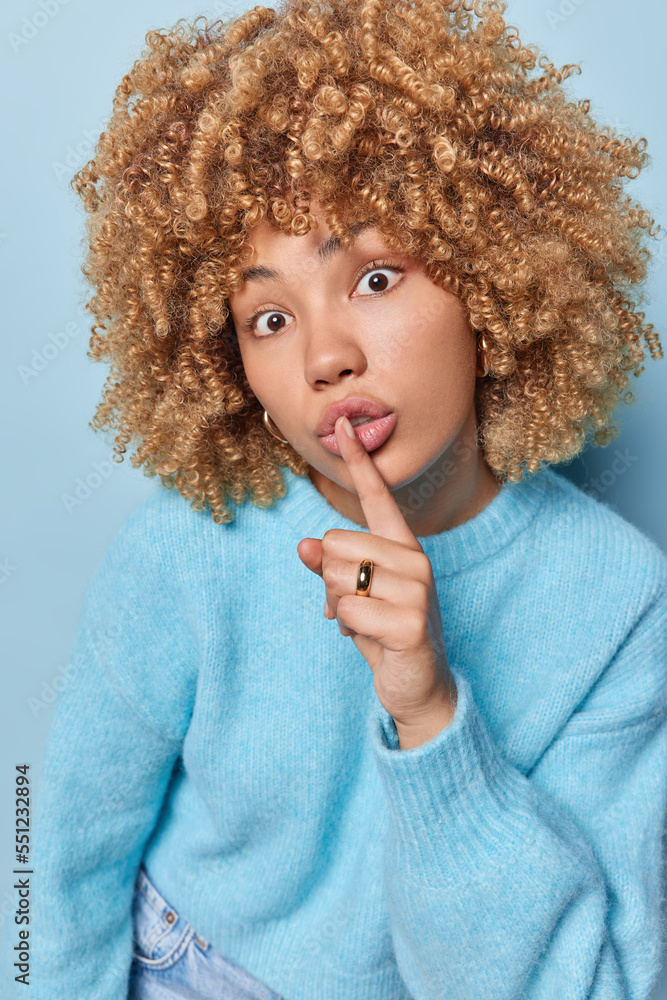 Vertical shot of surprised mysterious woman with curly hair makes silence gesture keeps index finger to lips tells secret dressed in casual jumper poses against blue background. People and secrecy Vertical shot of surprised mysterious woman with curly hair makes silence gesture keeps index finger to lips tells secret dressed in casual jumper poses against blue background. People and secrecy