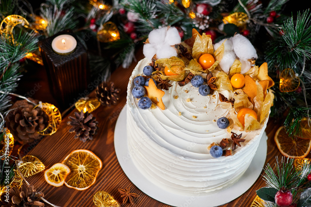 Christmas cake made of chocolate base with cream, decorated with blueberries and physalis against the backdrop of Christmas decor
