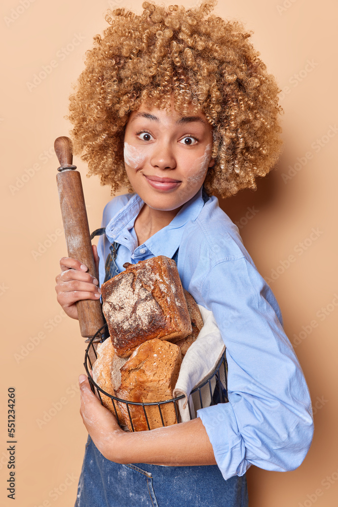 Surprised curly female baker holds rolling pin and basket offresh crusty bread enjoys baking organic yeast free products dressed in shirt and apron isolated over beige background. Bakery concept Surprised curly female baker holds rolling pin and basket offresh crusty bread enjoys baking organic yeast free products dressed in shirt and apron isolated over beige background. Bakery concept