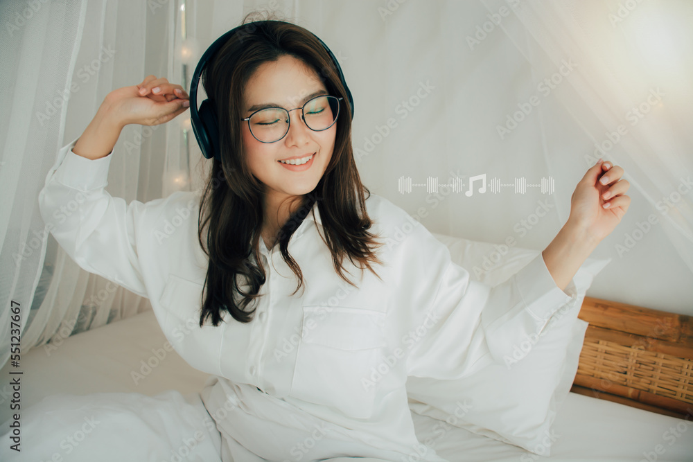 Funny young woman with beaming smile in eyewear and wearing headphones. Listening to music singing and dancing on bed at home. She wears white shirt. Music lover fan hobby concept. Music note icon. Funny young woman with beaming smile in eyewear and wearing headphones. Listening to music singing and dancing on bed at home. She wears white shirt. Music lover fan hobby concept. Music note icon.