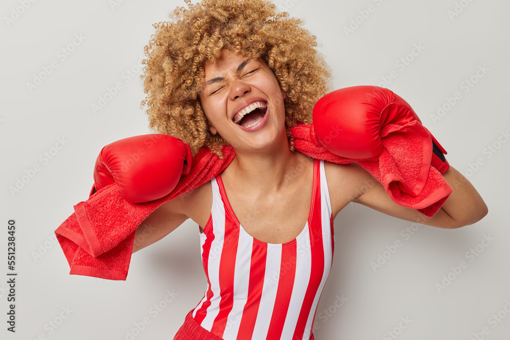 Overjoyed curly haired sportswoman enjoys boxing laughs positively wears striped t shirt protective boxing gloves prepares for final competition isolated over grey background. Sport and people concept