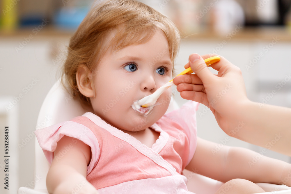 Very cute and funny baby with the spoon in hands eating kids nutrition - fruit puree. Smiling little girl eating in fun pose on white feeding table. Concept of first organic child food and family care