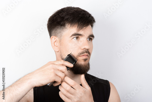 Bearded man trimming his beard with a trimmer on a light background